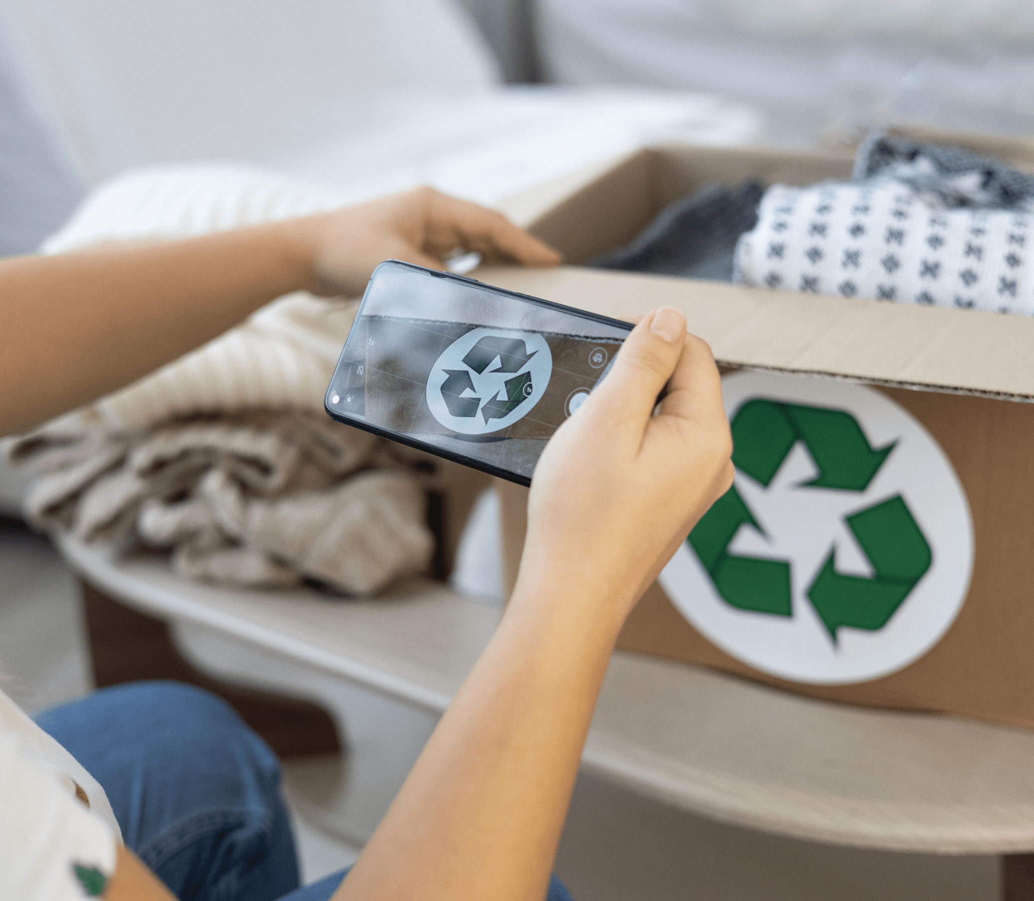 Person taking a photo of a cardboard box with a recycle symbol on it, surrounded by textiles, using a smartphone.