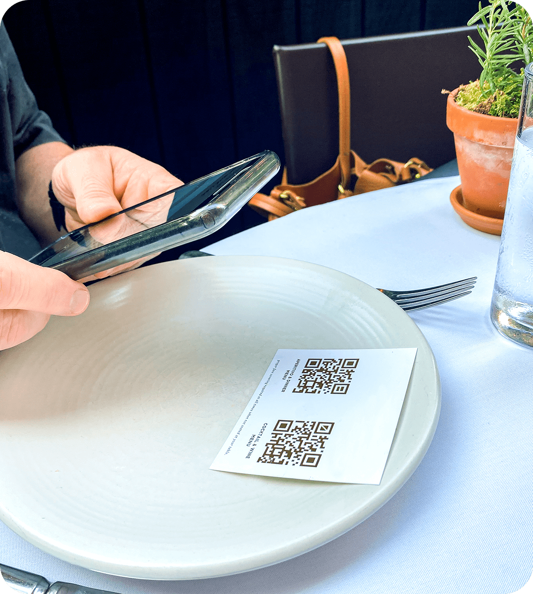 A person holding a smartphone scans a QR code on a card placed on a white plate at a restaurant table.